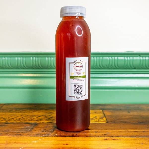 A bottle of Umami Global Bistro's Peach Ginger Chai on a wooden table with a green wall.