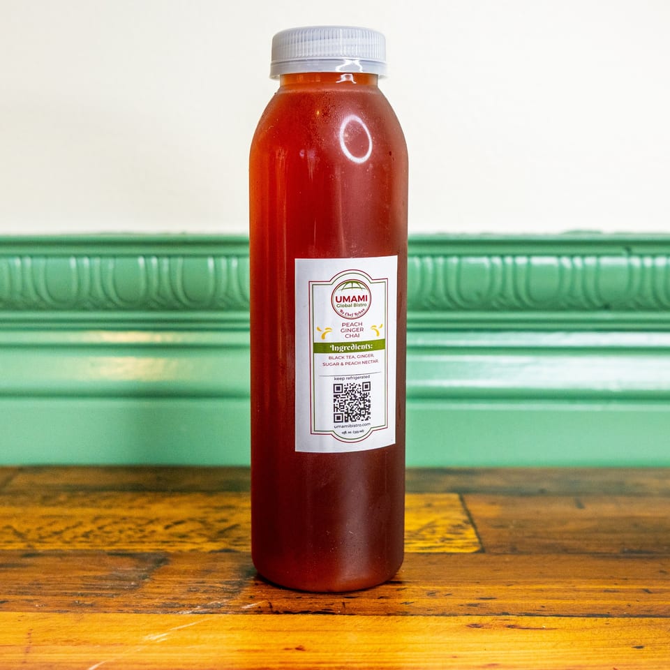 A bottle of Umami Global Bistro's Peach Ginger Chai on a wooden table with a green wall.