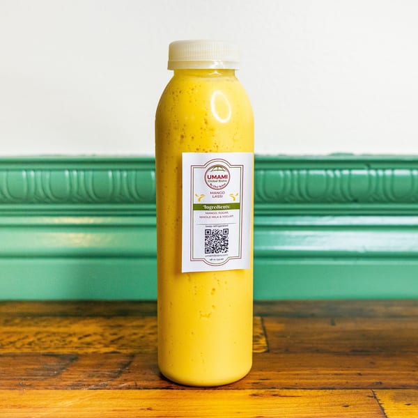 Umami Global Bistro's Mango Lassi drink in a clear bottle with a white label, on a wooden surface.